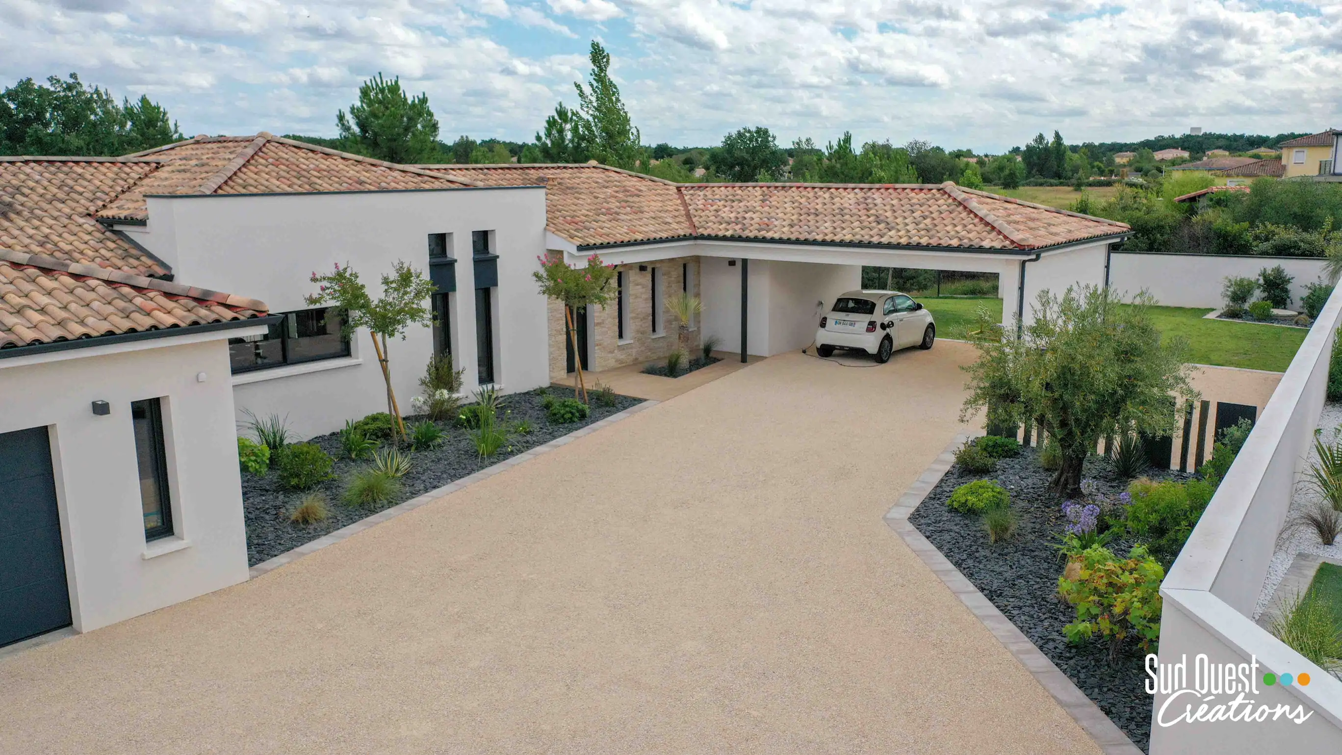 Cour carrossable en revêtement clair menant à un grand appentis attenant à la maison, utilisé comme carport pour une voiture électrique branchée.