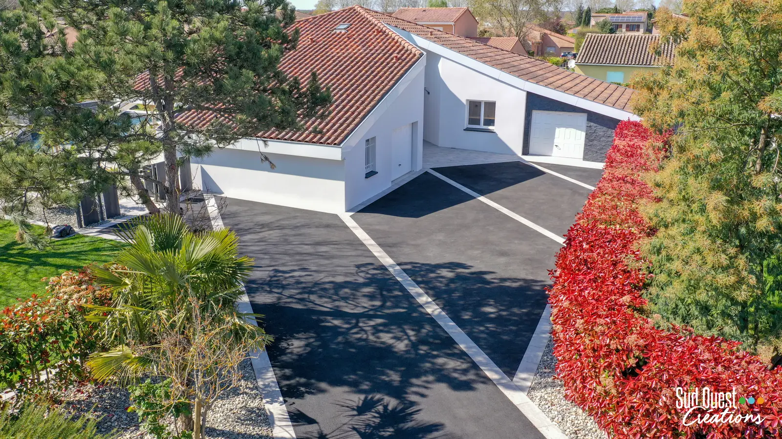 Allée carrossable en enrobé noir rythmée de bandes de pierre claire, bordée d'une haie de photinias