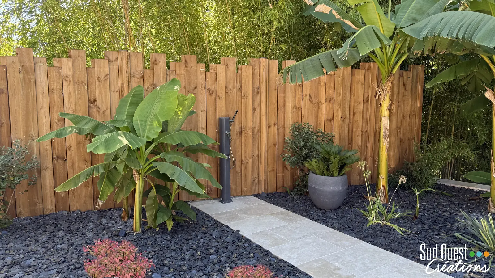 Douche extérieure entre bananiers et cycas devant une clôture en bois brut
