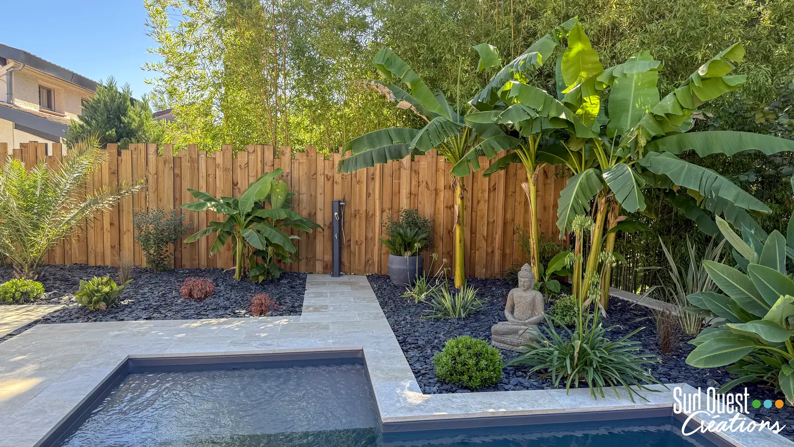 Douche de piscine au bout d'une allée en travertin, entre bananiers et clôture bois
