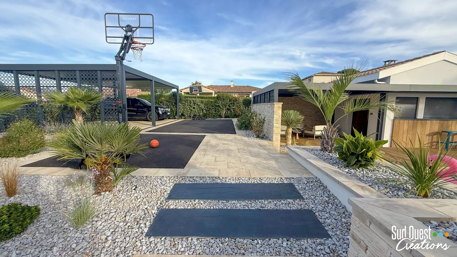 Entrée paysagée avec pas japonais sur galets blancs, palmiers et carport, terrain de basket