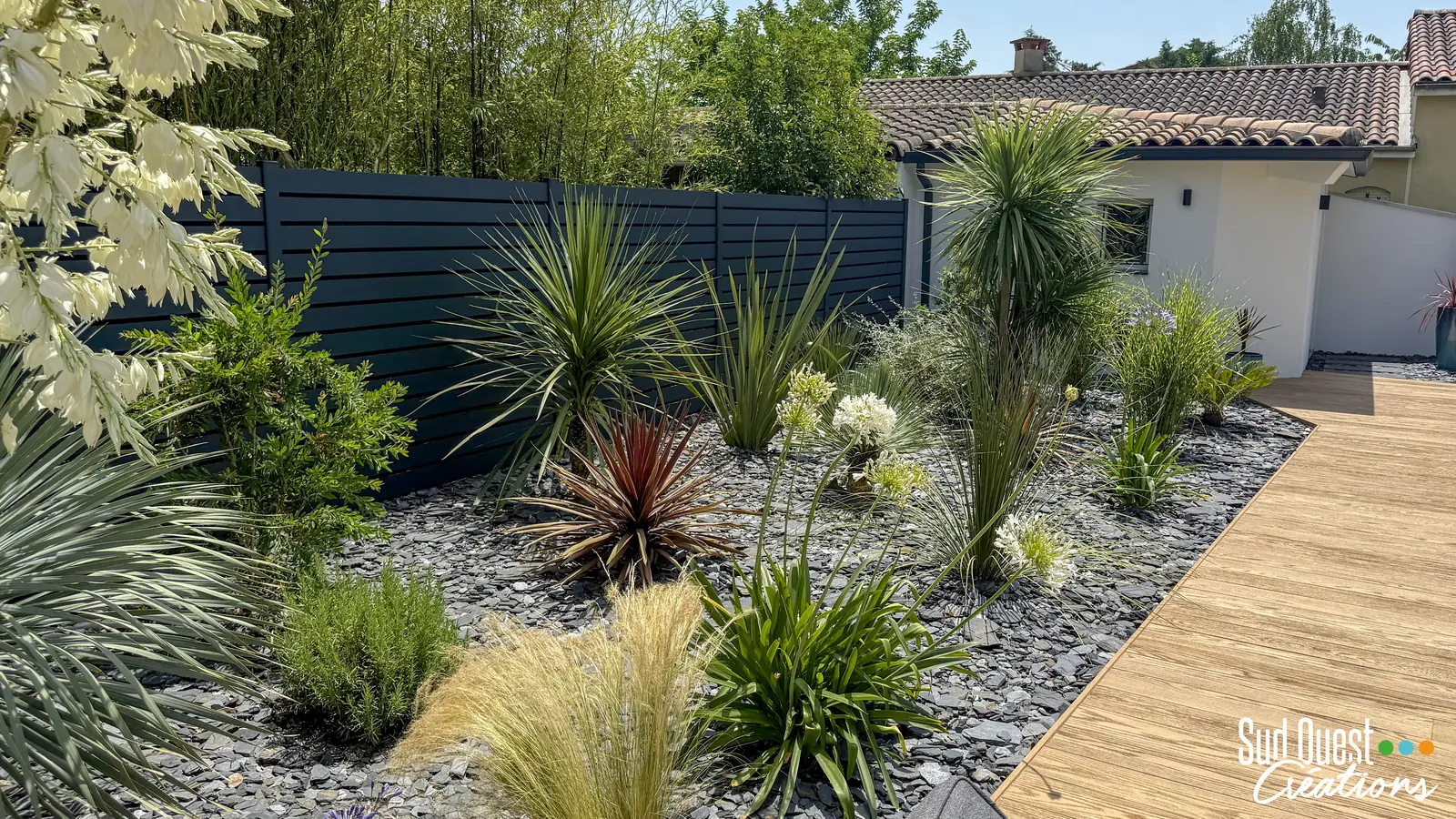 Massif sec sur ardoise avec yuccas, cordylines, agapanthes et graminées devant une clôture aluminium