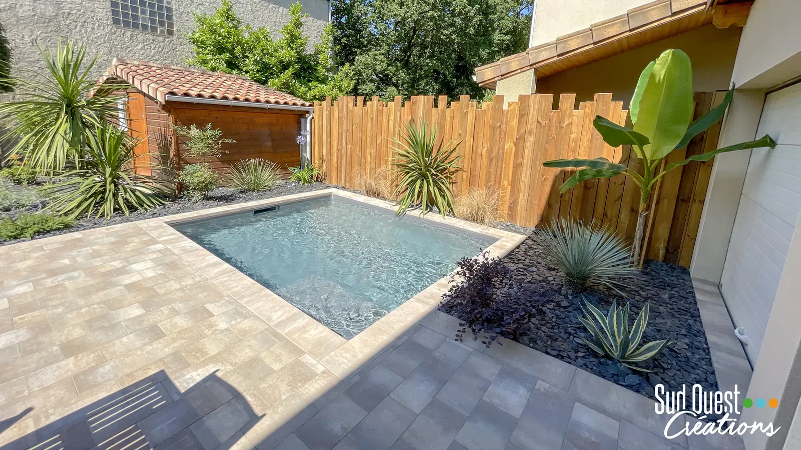 Petite piscine maçonnée dans une cour urbaine avec plage en pavés, clôture bois, bananier et paillage d'ardoise