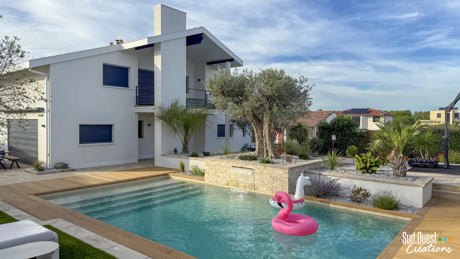 Piscine avec cascade sur mur en pierre, olivier centenaire et plage composite devant une maison contemporaine