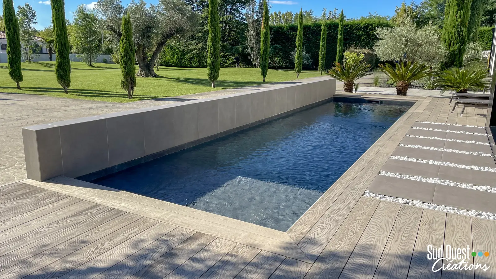 Piscine couloir au liner sombre avec mur habillé de grès cérame, terrasse composite et alignement de cyprès
