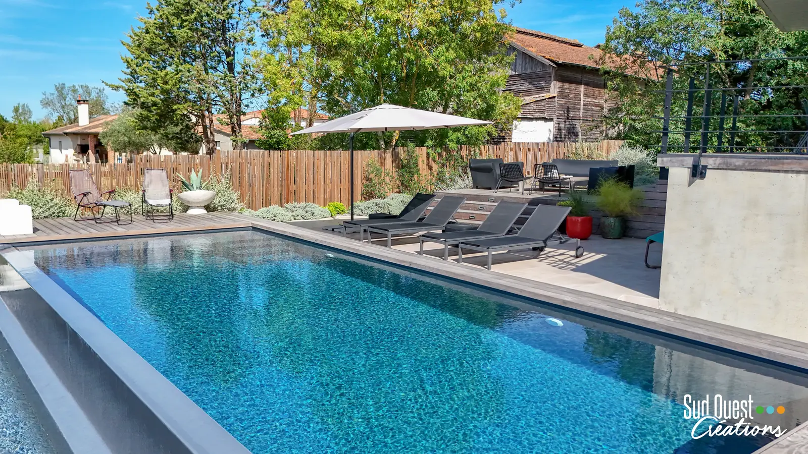 Piscine à débordement au liner gris, terrasse bois, clôture en bois brut et transats sous parasol