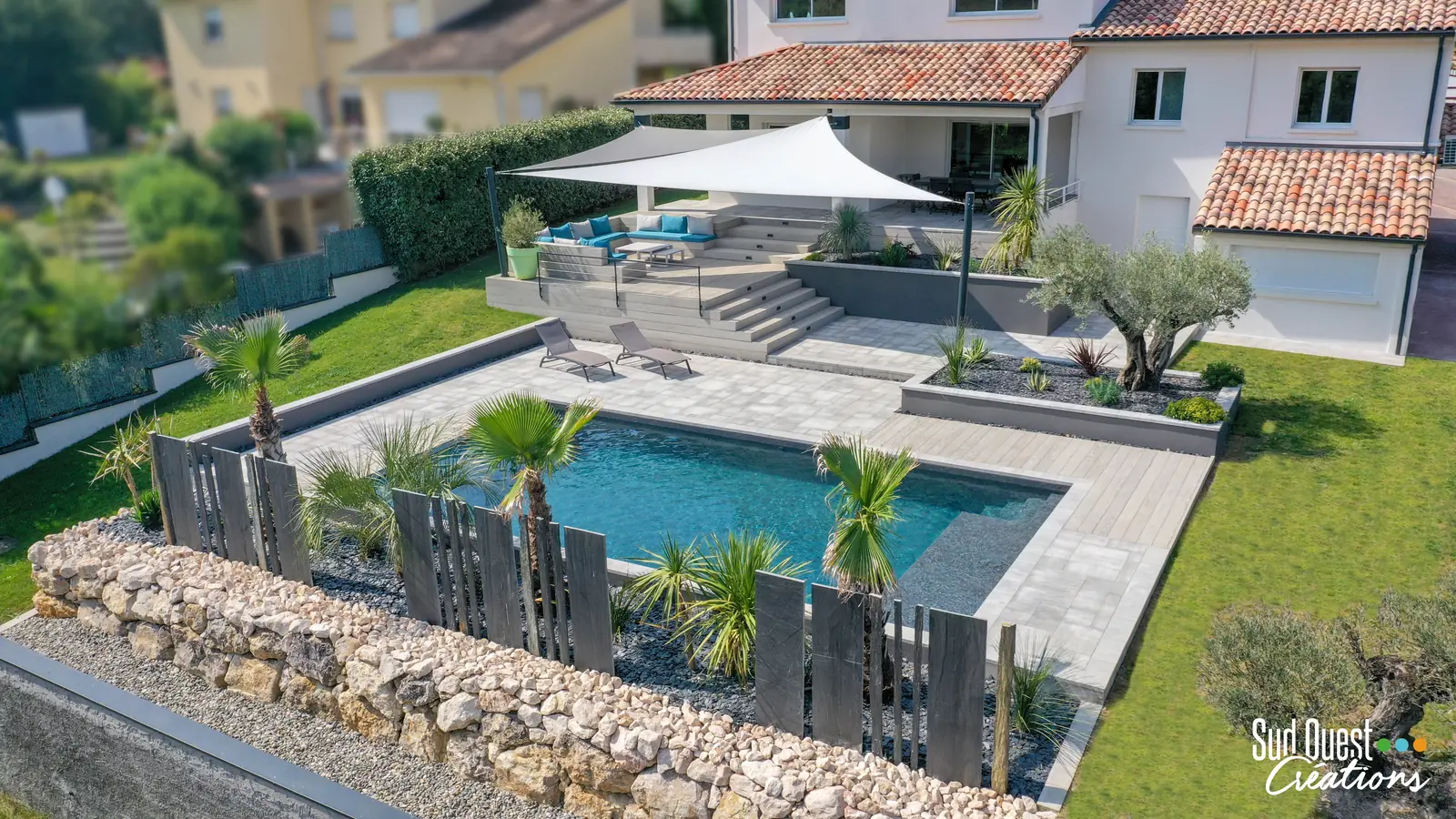 Vue drone d'une piscine bordée de palmiers, de lames d'ardoise dressées et d'un mur en pierre sèche