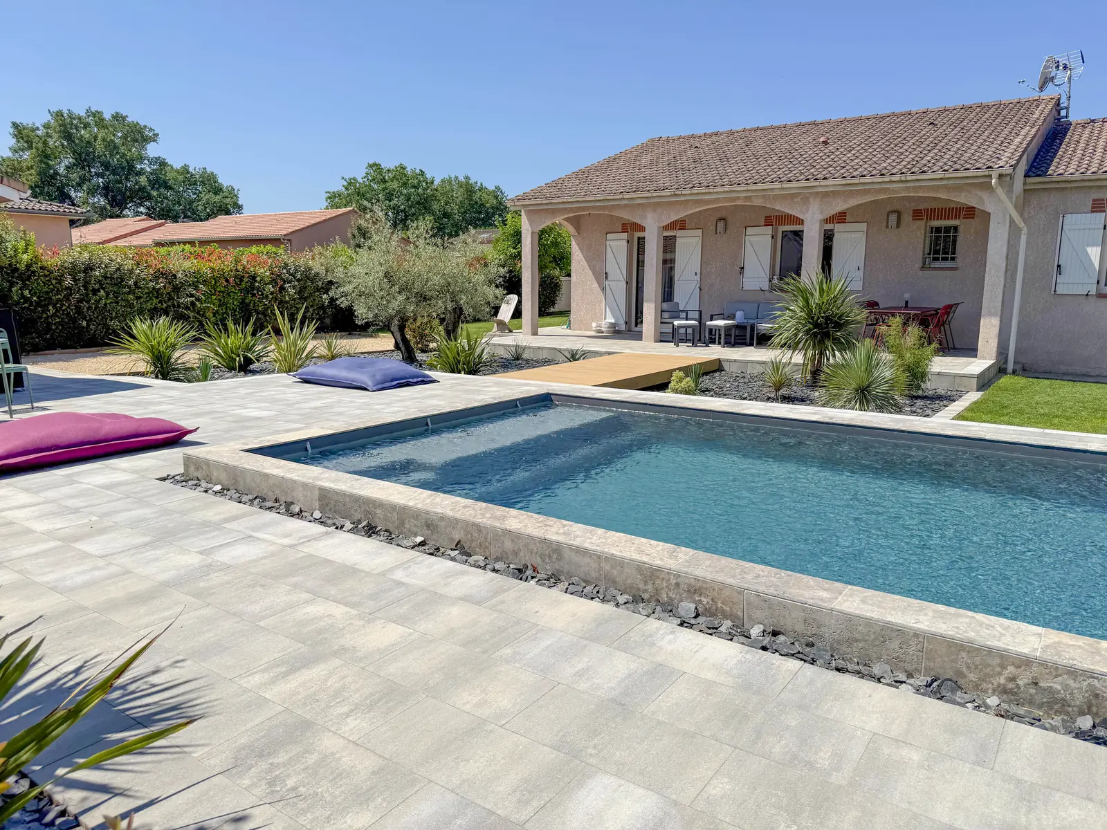 Piscine avec margelles en travertin, plage en dallage gris, olivier et passerelle en bois