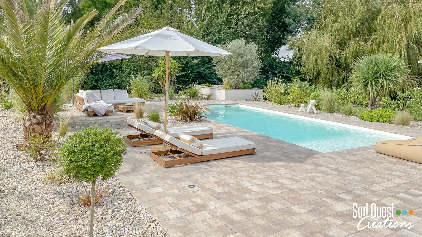 Piscine avec plage en pavés dans un jardin exotique de palmiers, graminées et galets