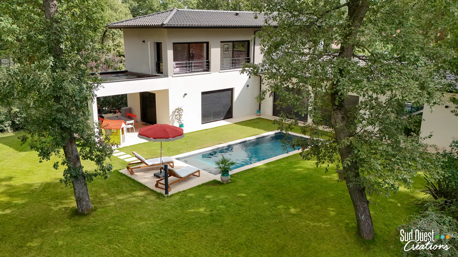Vue drone d'une piscine rectangulaire devant une maison contemporaine, pelouse et chênes