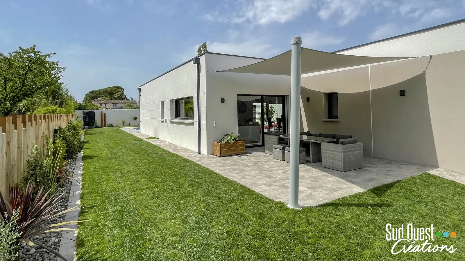 Voile d'ombrage sur terrasse en pavés devant une maison contemporaine, pelouse et clôture bois